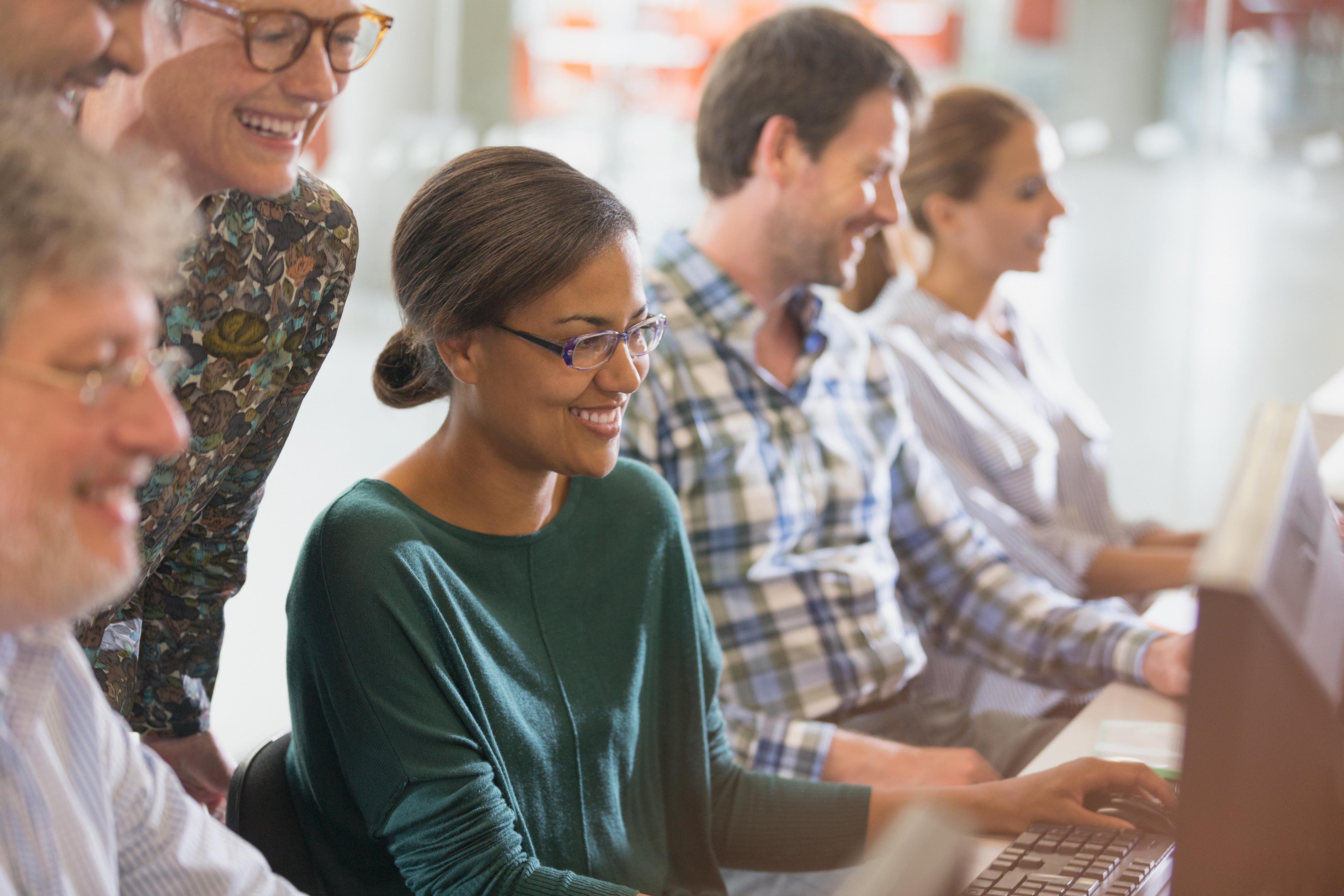 Das Bild zeigt eine Gruppe von Erwachsenen, die lächelnd an Computern in einem hellen Raum sitzen und zusammenarbeiten.  Im Detail zeigt das Bild mehrere Erwachsene, die an Computern sitzen, wahrscheinlich in einem Computerlabor oder einer ähnlichen Umgebung. Im Vordergrund steht eine junge Frau mit dunkler Hautfarbe und Brille, die konzentriert an einem Computer arbeitet. Sie trägt ein dunkelgrünes Langarmshirt und lächelt leicht. Hinter ihr und leicht über ihre Schulter gebeugt, stehen zwei ältere Frauen, die ebenfalls lächeln. Eine der Frauen trägt eine bunt gemusterte Bluse. Links im Bild ist ein älterer Mann mit Brille zu sehen, dessen Gesicht teilweise verdeckt ist. Er lächelt ebenfalls. Neben der jungen Frau im Vordergrund sitzt ein Mann mit kariertem Hemd, der ebenfalls mit einem Computer arbeitet und lächelt. Im Hintergrund sind weitere Personen zu sehen, die ebenfalls an Computern arbeiten, sie sind jedoch unscharf und weniger detailliert erkennbar. Die allgemeine Atmosphäre des Bildes ist freundlich, kollaborativ und positiv. Die warme Beleuchtung und die lächelnden Gesichter der Personen tragen zu einer angenehmen und entspannten Stimmung bei. Das Bild vermittelt den Eindruck von gemeinsamer Arbeit, Lernen oder Weiterbildung in einer unterstützenden Umgebung.