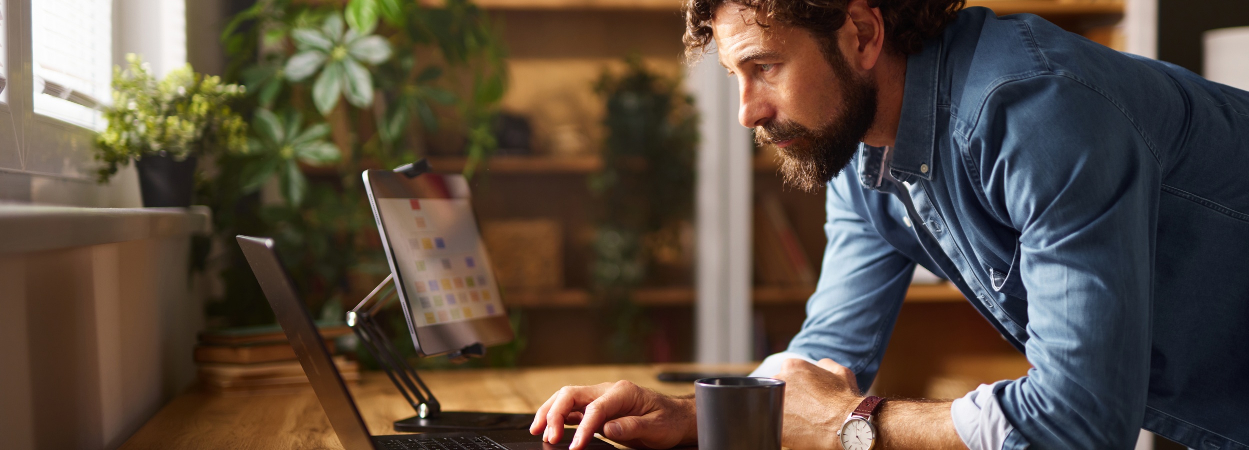 Mann mit Jeanshemd arbeitet konzentriert im Büro an seinem Laptop. Er beugt sich dafür auf den Schreibtisch und stützt sich mit den Armen auf.