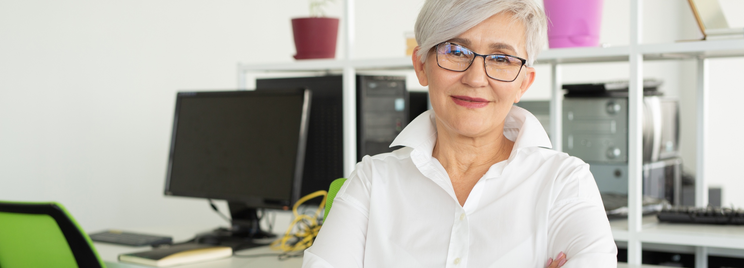 Ältere Arbeitnehmerin mit grauen Haaren und weißer Bluse arbeitet in einem Büro.