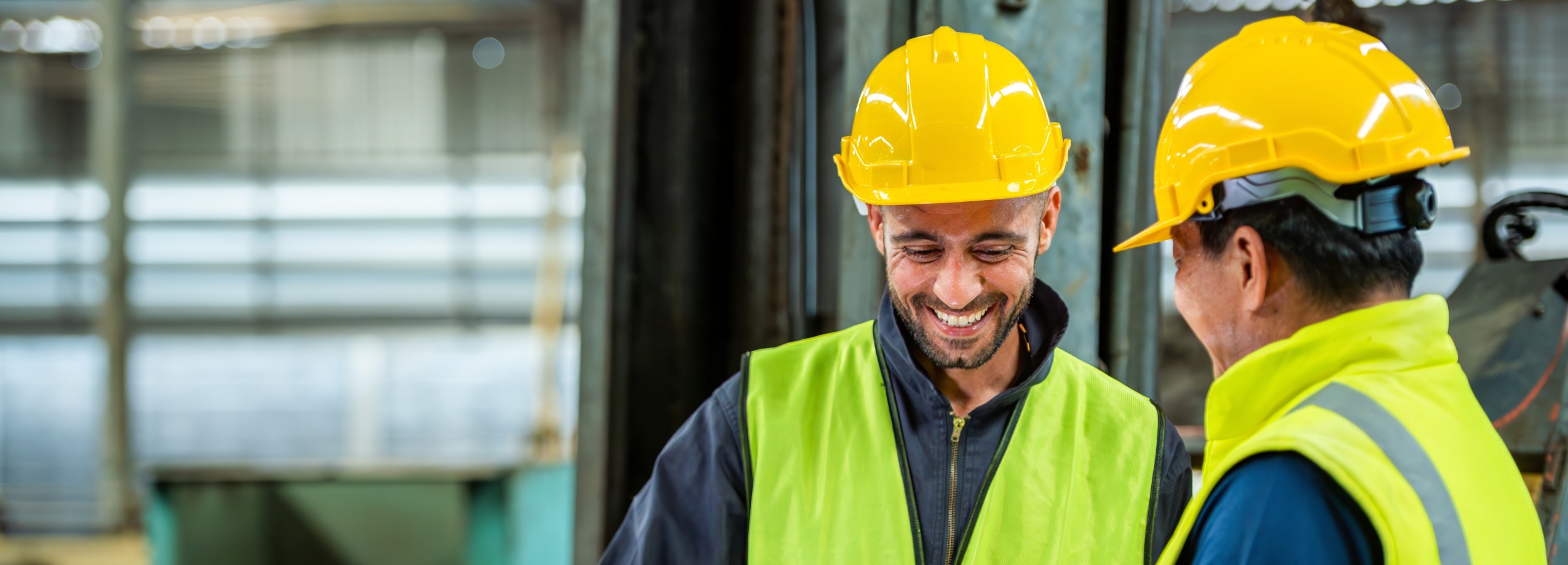 Zwei Personen tragen gelbe Schutzhelme und neongelbe Warnwesten. Sie stehen in einer Industriehalle mit großen Fenstern und sprechen miteinander.
